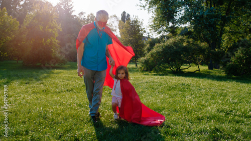 In front of the camera old man and his cute little niece wearing a superhero suit walking in front of the camera in the middle of the park