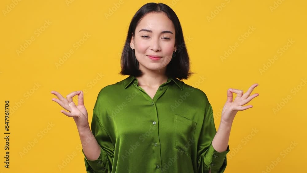 Beautiful spiritual tranquil young woman of Asian ethnicity she wear green shirt spreading hands in yoga om aum gesture relax meditate try to calm down isolated on plain yellow color wall background Beautiful spiritual tranquil young woman of Asian ethnicity she wear green shirt spreading hands in yoga om aum gesture relax meditate try to calm down isolated on plain yellow color wall background