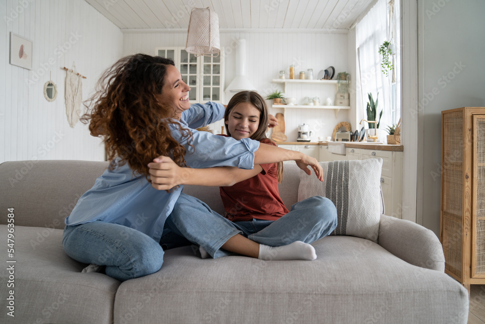 Cheerful friendly family of woman and teenage girl want to hug after short separation. Positive smiling Caucasian mother sits on sofa in home interior and enjoys communicating with beloved daughter 