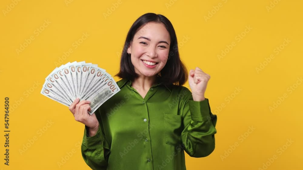 Young woman of Asian ethnicity 20s she wear green shirt hold in hand fan of cash money in dollar banknotes doing winner gesture celebrate isolated on plain yellow color wall background studio portrait Young woman of Asian ethnicity 20s she wear green shirt hold in hand fan of cash money in dollar banknotes doing winner gesture celebrate isolated on plain yellow color wall background studio portrait