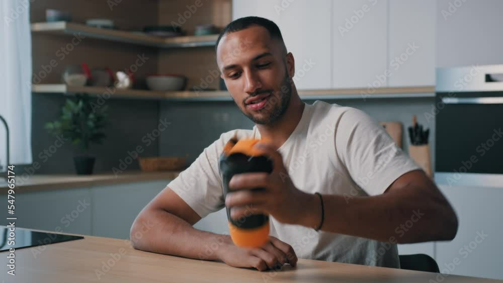 African American sport man male athlete trainer bodybuilder sitting at kitchen table shaking bottle shaker with liquid calcium mixing protein cocktail sports supplements healthy nutrition fitness diet
