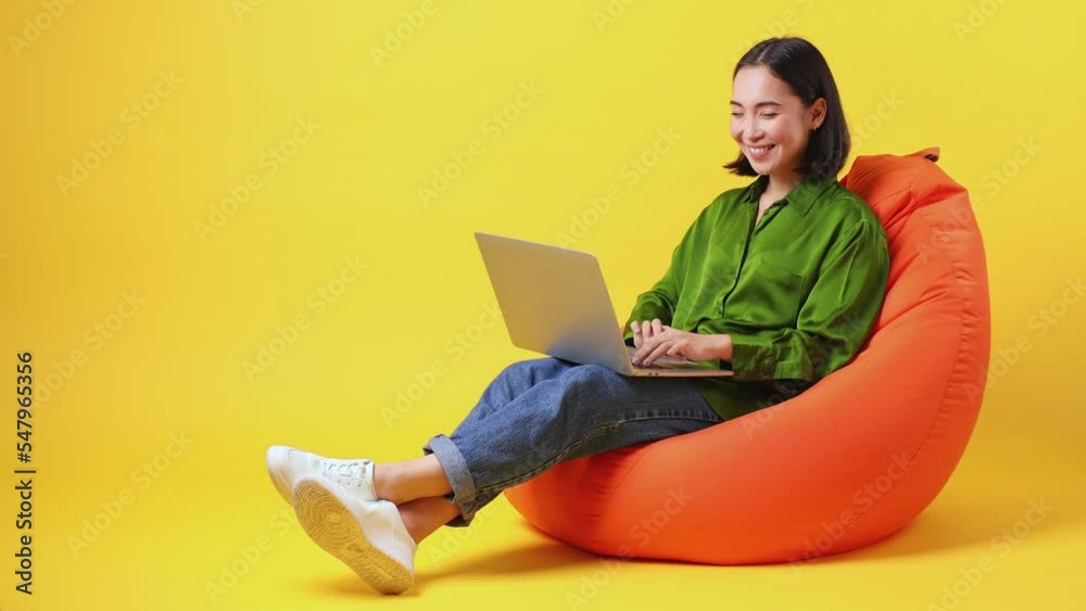Full size body length smiling IT young woman of Asian ethnicity she wear green shirt sit in bag chair hold use work on laptop pc computer isolated on plain yellow color wall background studio portrait