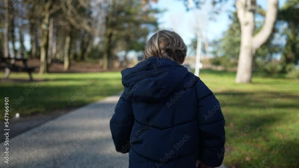 Back of child walking outside in park. One small boy wearing jacket strolling outdoors during sunny day in winter season Back of child walking outside in park. One small boy wearing jacket strolling outdoors during sunny day in winter season