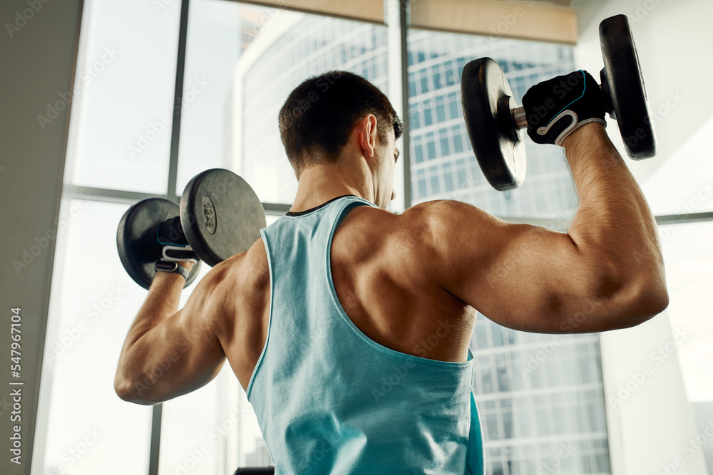 Man doing exercise with dumbbell leaning on sports bench in the gym. Photo of a sexy muscular man in sportswear and good physique on grey background. Strength and motivation, sport, fitness goal Man doing exercise with dumbbell leaning on sports bench in the gym. Photo of a sexy muscular man in sportswear and good physique on grey background. Strength and motivation, sport, fitness goal