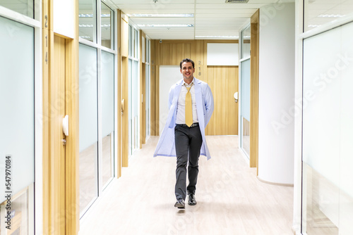 Determined handsome doctor or medic wearing white coat walking along hospital hallway. Modern bright clinic © TeTe Song