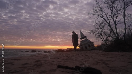 Woman walking from mysterious old flooded leaning wooden house, chapel washed by sea waves, sinking in water on beach. Girl goes away after visiting damaged abandoned home on vivid dreamy pink sunset. photo