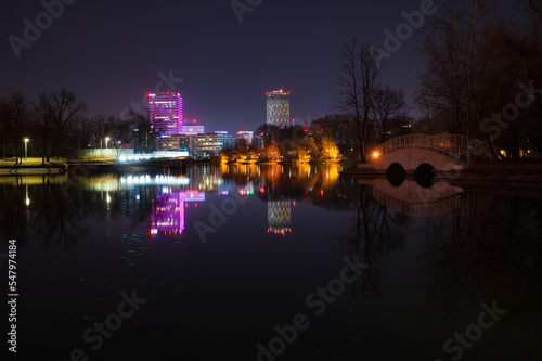 Obraz na płótnie Beleuchtung im Parcul Bordei in Bukarest, mit Fußgängerbrücke und modernen Bürotürmen um den See im Hintergrund w salonie