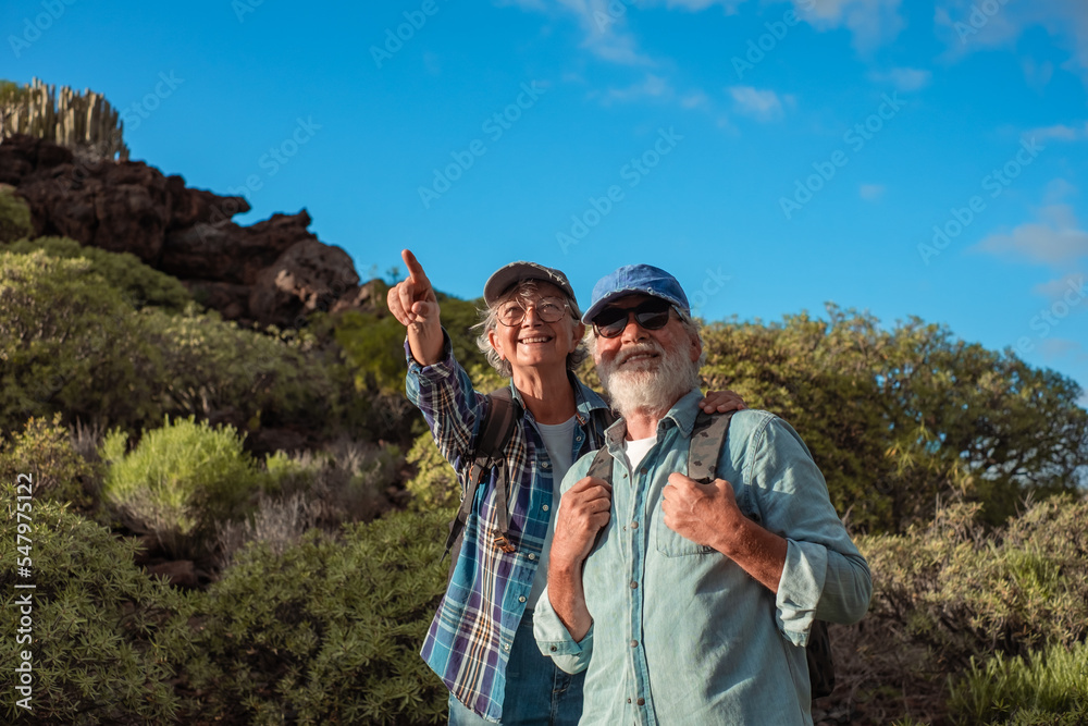 Happy senior couple in mountain trekking enjoying nature, freedom and healthy lifestyle. Smiling old retirees in hat and casual clothes among green bushes and blue sky