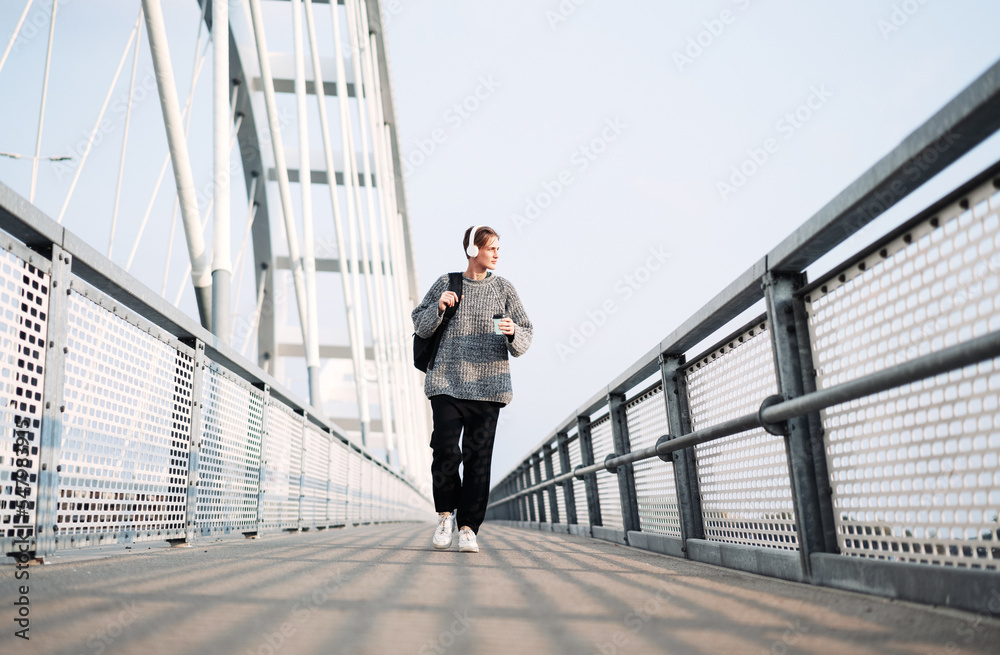 Confident young woman listening music with her headohones while walking across the footbridge.