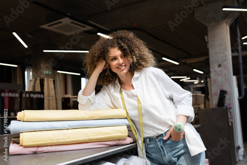 cheerful saleswoman with needle cushion posing with hand in pocket while leaning on desk with colorful fabric rolls