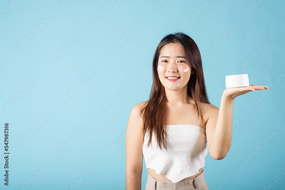 Asian young woman are not very beautiful hold jar of skin cream for face and apply anti-aging moisturizer studio isolated on blue background, Happy female pointing finger her face, pursuit of beauty Asian young woman are not very beautiful hold jar of skin cream for face and apply anti-aging moisturizer studio isolated on blue background, Happy female pointing finger her face, pursuit of beauty