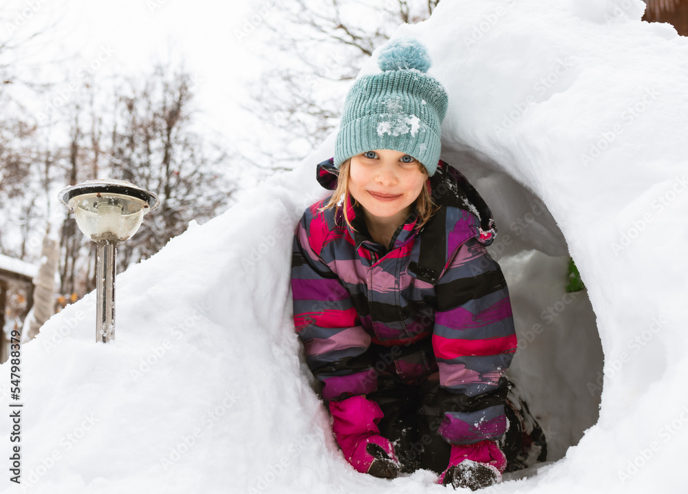 Fototapeta premium Happy little girl sitting in a snow cave made in a snowdrift looking at camera smiling