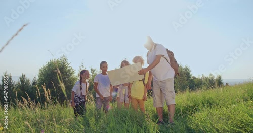 Front view of children standing together looking for route with old man holding map, showing, explaining. Tourists traveling in hills, hiking. Concept of tourism and team spirit. 