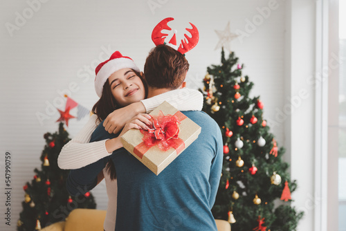 Asian young couple in love hugging each other in living room decorated Christmas tree, exchanging Christmas presents and celebrating New Year