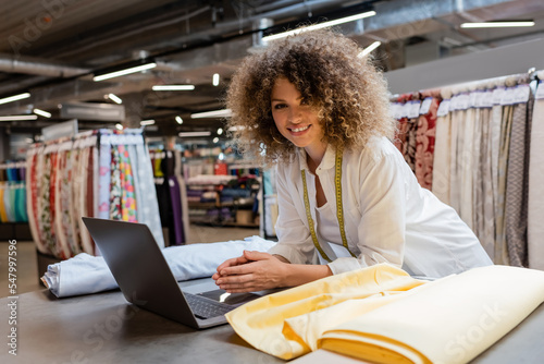 joyful saleswoman with curly hair using laptop near fabric rolls in textile shop