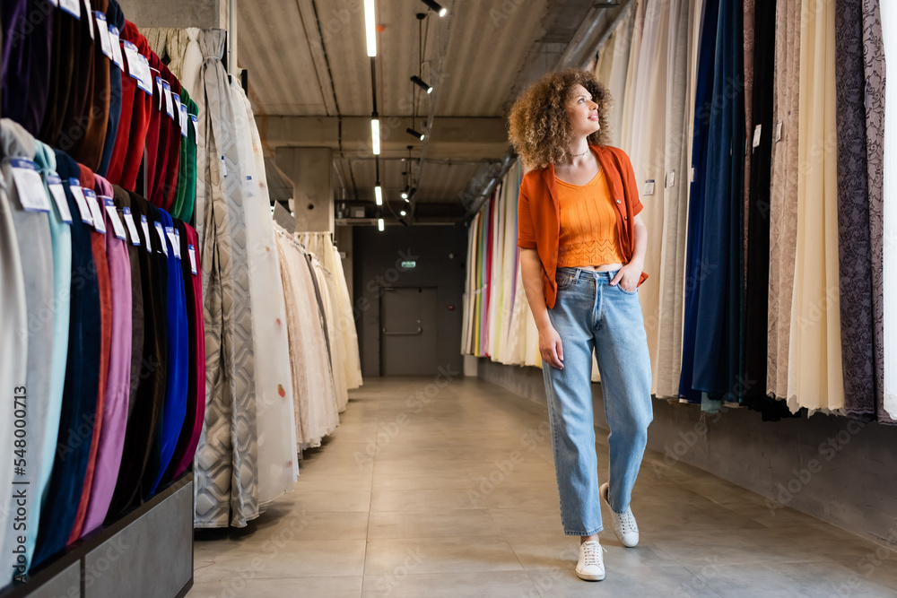 full length of curly woman holding hand in pocket of jeans and looking at curtains in textile shop