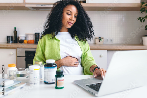 Picture of adorable african american pregnant woman ordering prenatal supplements in web store sitting at kitchen table with bottles of vitamins, scrolling on laptop. Healthy pregnancy photo