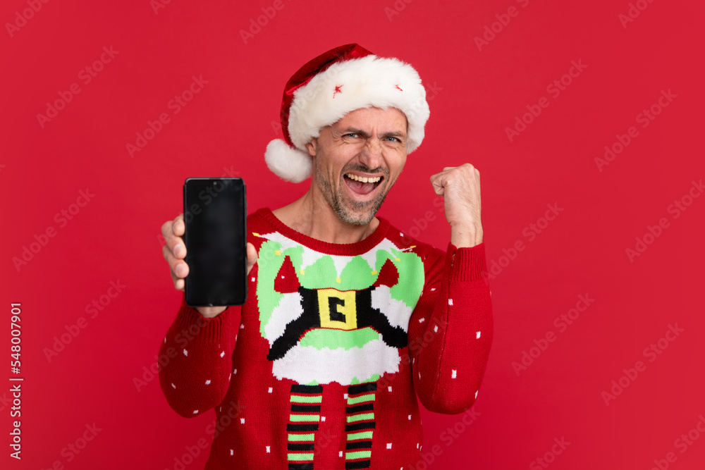 Santa with phone screen. Man in Christmas sweater and hat on color background. Middle aged santa.