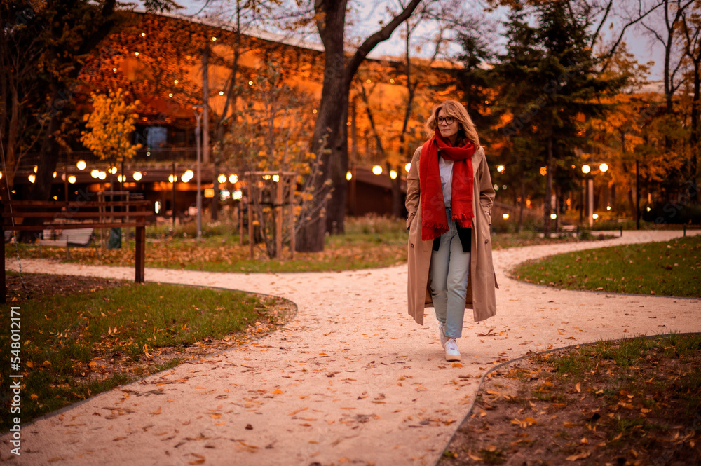 Fashionable middle aged woman walking in the street and enjoying autumn afternoon. Attractive female wearing scarf and trench coat outdoor. Fashionable middle aged woman walking in the street and enjoying autumn afternoon. Attractive female wearing scarf and trench coat outdoor.
