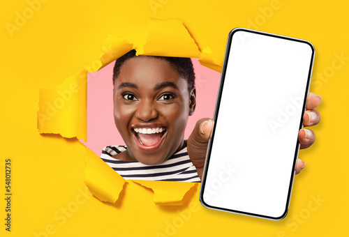 Excited black lady showing phone through torn yellow paper hole © Prostock-studio