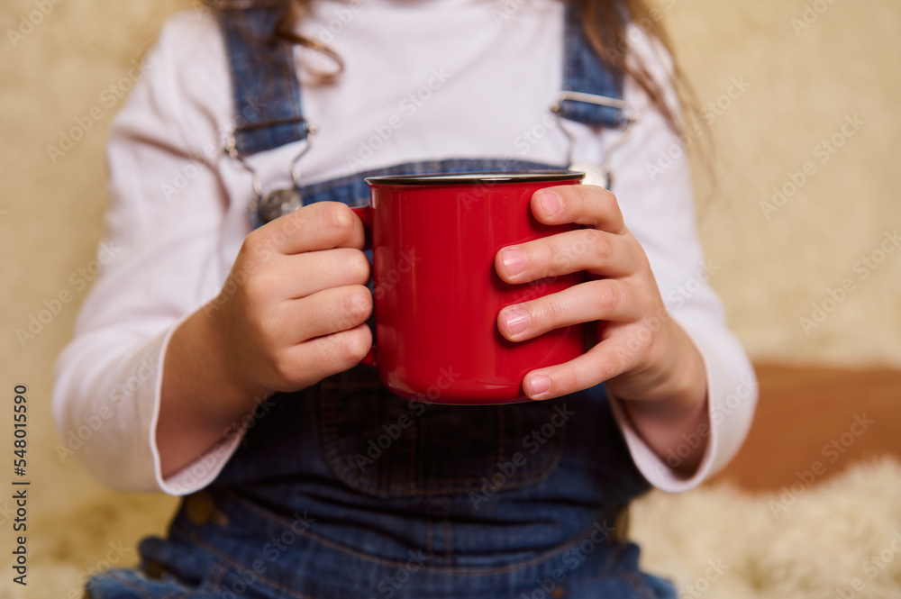 Selective focus on a red mug of hot cocoa or chocolate drink with marshmallows in the hands of a little child girl, enjoying happy festive atmosphere of Christmas winter holidays at home interior