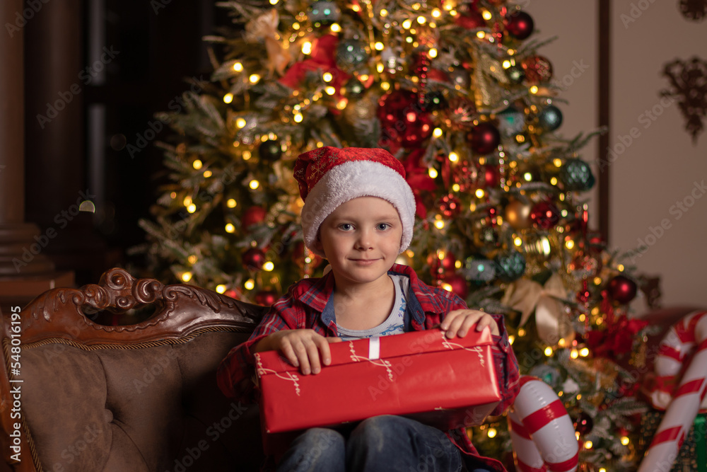 Adorable little kid in a santa hat with gift box in his hand on the background of the christmas tree