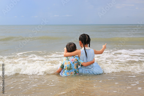 Back view of Asian young girl child hugging the neck little brother sitting on tropical sand beach at sunrise. Adorable sister and brother having fun in summer holiday.