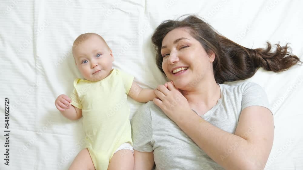 Happy mother and newborn baby lie on bed looking in camera and smiling. Young woman enjoys resting at home with little child upper close view Happy mother and newborn baby lie on bed looking in camera and smiling. Young woman enjoys resting at home with little child upper close view