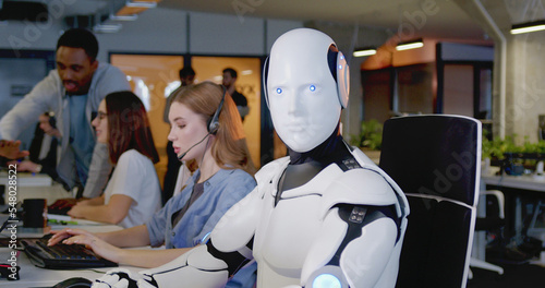 Digital transformation. Robot looking at camera in office. White device turning head and looking straight. Humanoid working at computer. Support worker. Artificial intelligence. Communication network.
