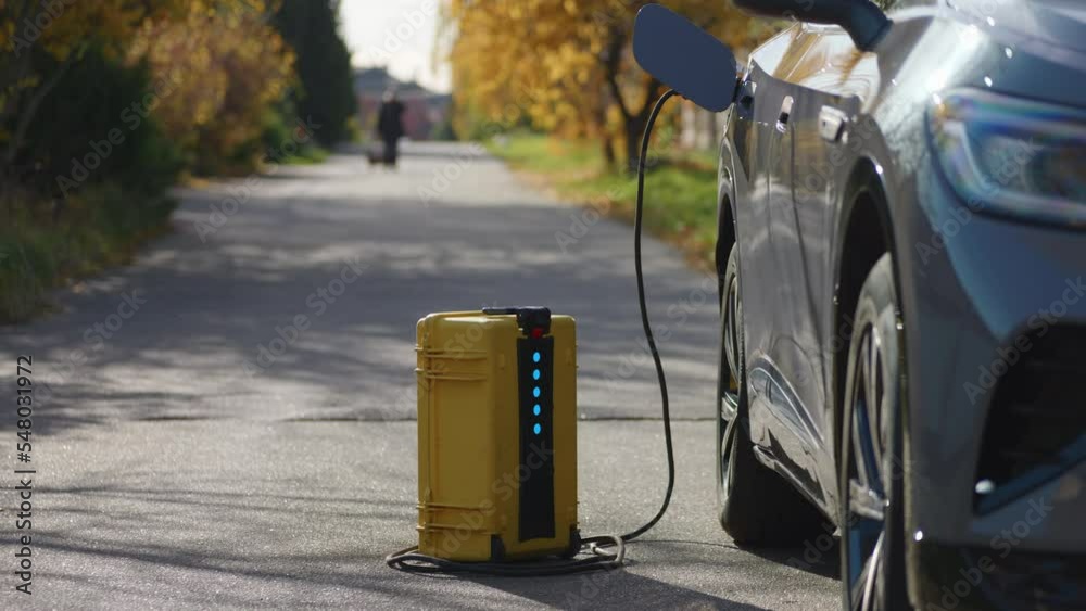 Using of home-invented portable battery to charge electric car out of the city. Light blinks indicating its work. Peaceful sunny autumn day, bright yellow leaves and road background. High quality 4k Using of home-invented portable battery to charge electric car out of the city. Light blinks indicating its work. Peaceful sunny autumn day, bright yellow leaves and road background. High quality 4k