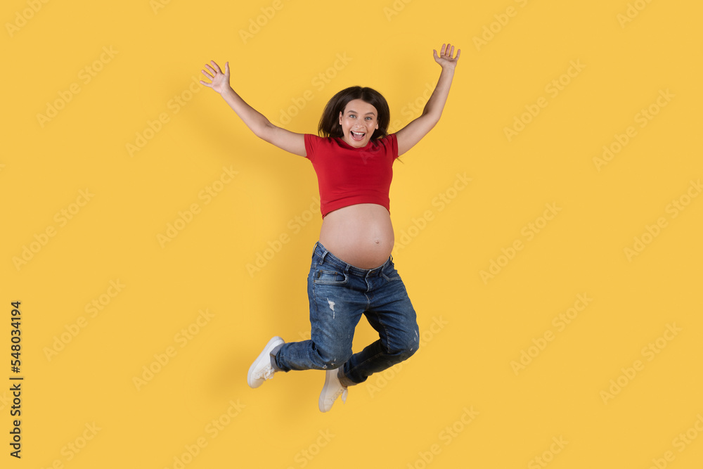 Cheerful Young Pregnant Woman Jumping With Raised Hands Over Yellow Background