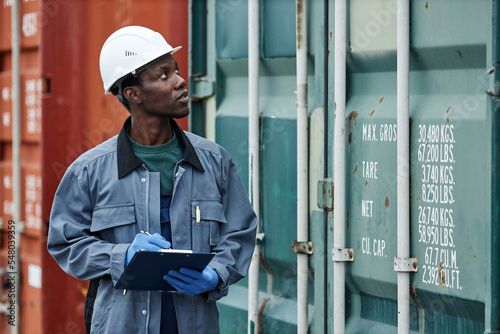 Waist up portrait of young black man wearing hardhat while working at shipping dock and holding clipboard photo