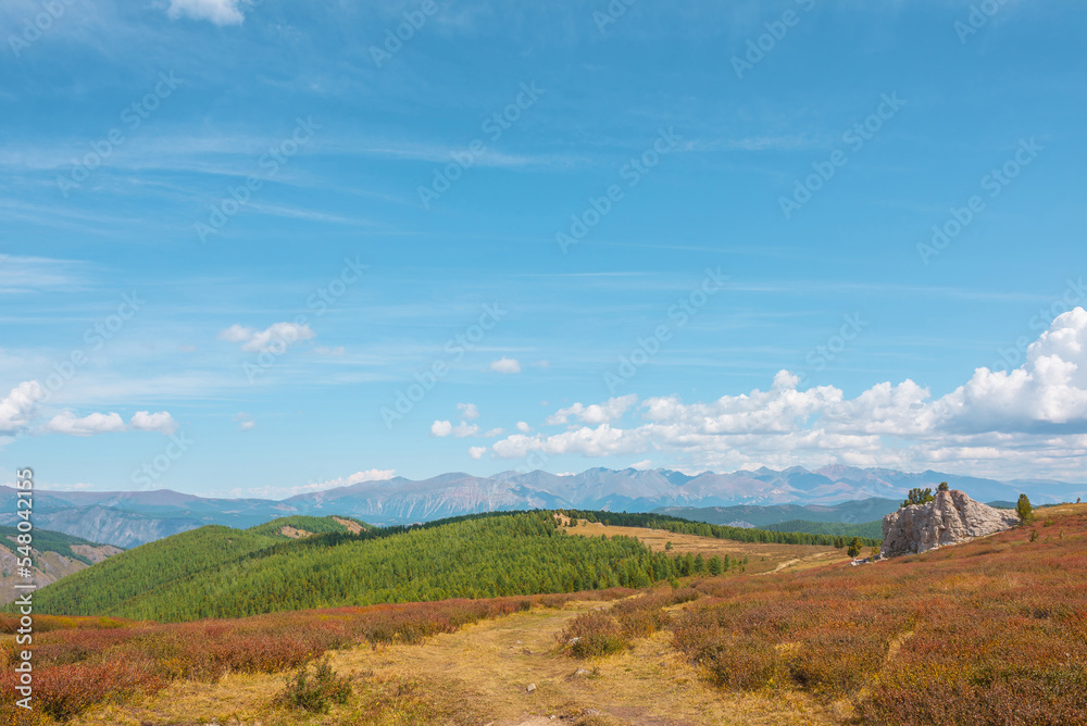 Atmospheric autumn landscape with big rock on hill with view to sunlit mountain vastness and high mountain range line on horizon under clouds. Impressive mountain scenery in vivid fading autumn colors