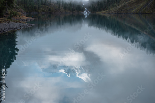 Tranquil scenery with reflex in mountain lake of snow castle in clouds. Snowy mountains in fog clearance  small river and coniferous trees reflected in alpine lake. Mountain creek and glacial lake.