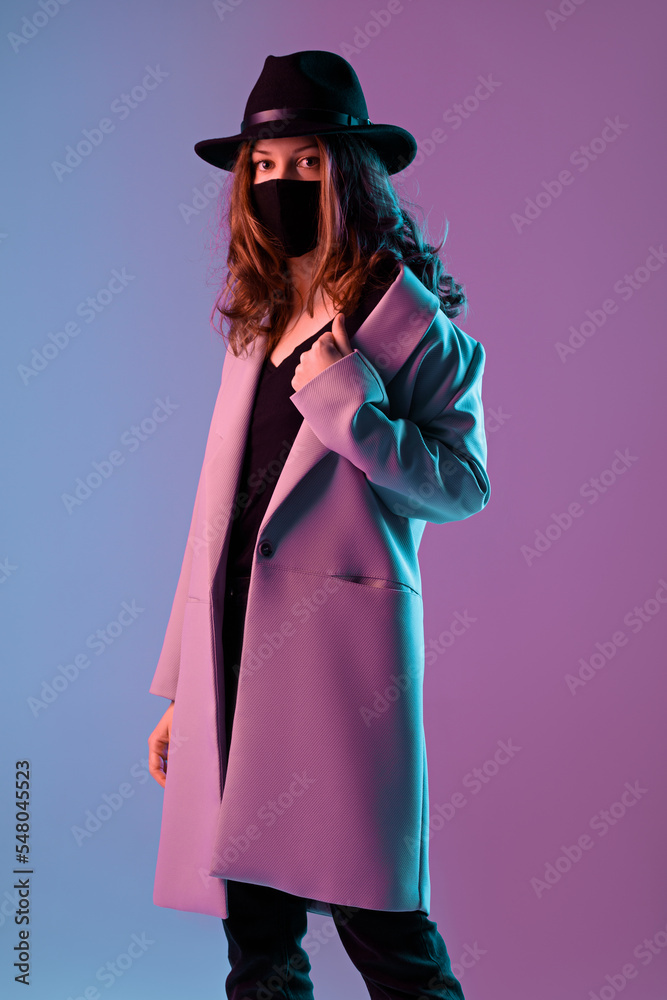 Portrait of a trendy young woman in a coat, hat and protective mask in purple neon lighting.