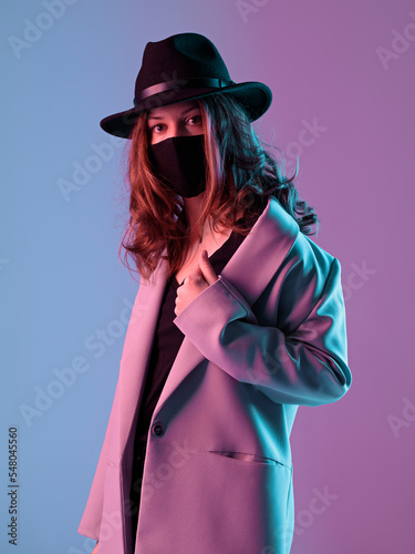 Obraz na płótnie Portrait of a fashionable young woman in a coat, hat and protective mask in purple neon lighting. w salonie
