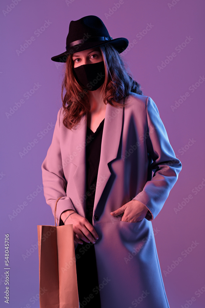 Portrait of a trendy young woman in a coat, hat and protective mask with a paper shopping bag in purple neon lighting.