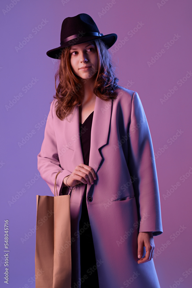 Portrait of a trendy young woman in a coat and hat with a paper shopping bag in purple neon lighting.