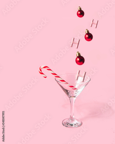 Hohoho Christmas cocktail. Candy cane, red bauble and glass on pastel light pink background. Minimalistic winter holidays greeting card. Creative New Year concept.
