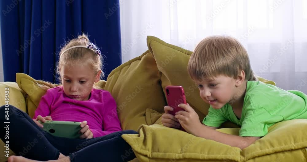 Excited preschooler girl and toddler boy enjoy playing games on smartphones at home. Siblings spending weekend on cosy sofa in apartment together