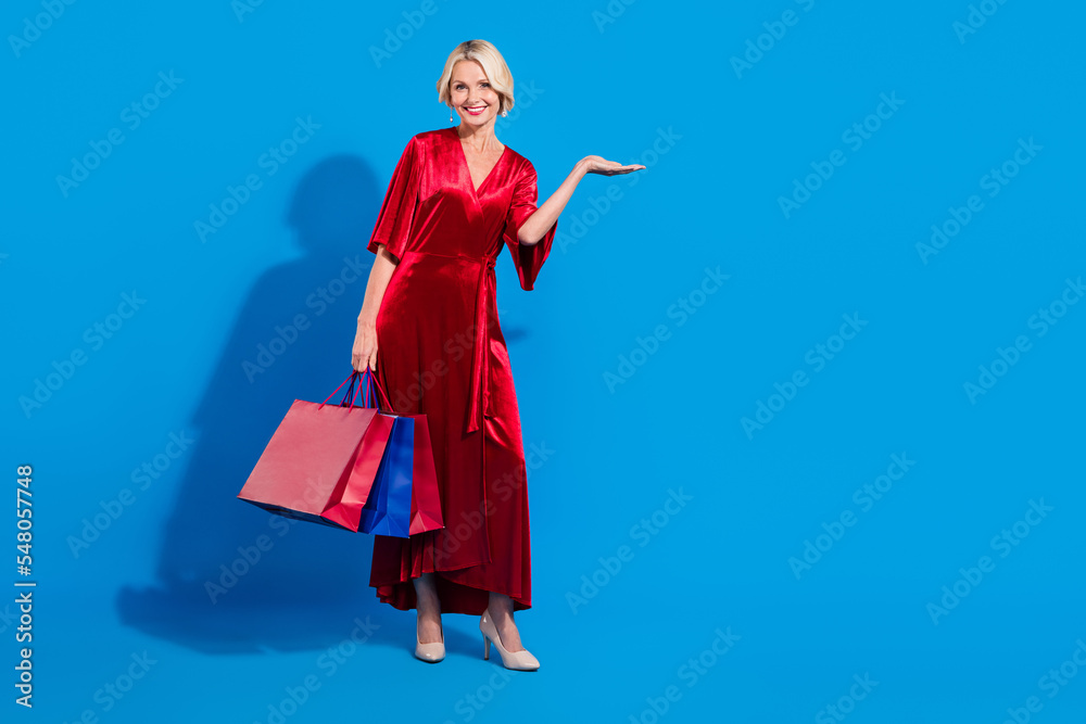 Full length photo of dreamy sweet retired lady dressed shiny red clothes holding shoppers arm empty space isolated blue color background Full length photo of dreamy sweet retired lady dressed shiny red clothes holding shoppers arm empty space isolated blue color background