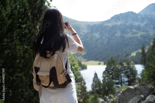 Mujer con mochila de excursión haciendo una foto con el móvil a un paisaje: lago en la montaña rodeado de árboles (Pirineo Catalán, Boi-Taull, Cataluña, España) photo