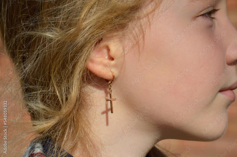 a gold earring in the shape of a cross in the ear of a young girl. fine gold jewelry for women of any age. beautiful girl. portrait in profile