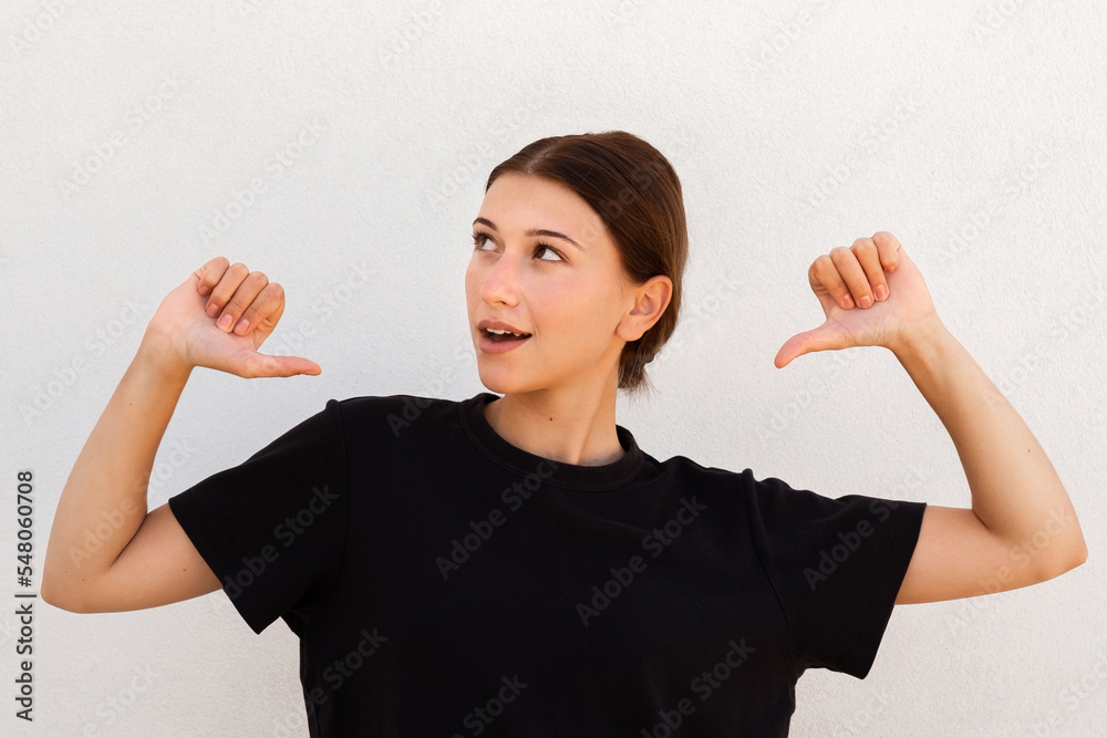 Portrait of happy young woman pointing thumbs at herself. Caucasian woman wearing black T-shirt looking away and smiling against white background. Pride concept Portrait of happy young woman pointing thumbs at herself. Caucasian woman wearing black T-shirt looking away and smiling against white background. Pride concept