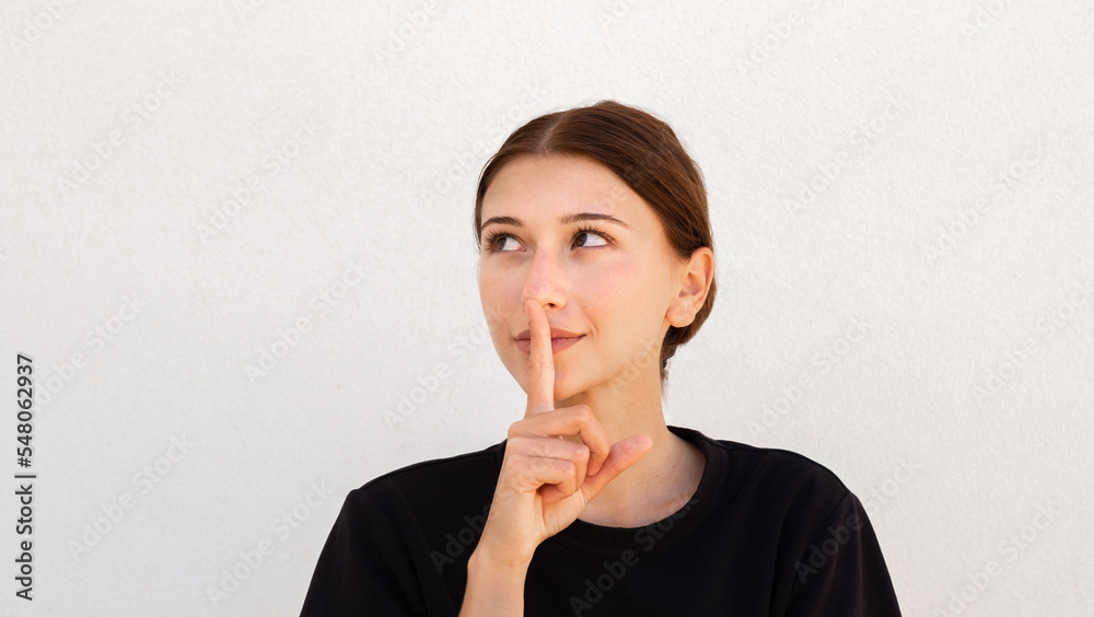 Portrait of confident young woman posing with finger on lips over white background. Caucasian woman wearing black T-shirt having secret. Silence and secrecy concept Portrait of confident young woman posing with finger on lips over white background. Caucasian woman wearing black T-shirt having secret. Silence and secrecy concept
