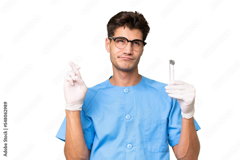 Young dentist man holding tools over isolated background with fingers crossing and wishing the best Young dentist man holding tools over isolated background with fingers crossing and wishing the best