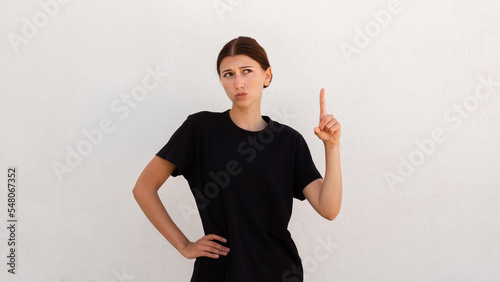 Portrait of doubtful young woman thinking over idea. Caucasian woman wearing black T-shirt pointing finger up with uncertain expression. Idea and solution concept