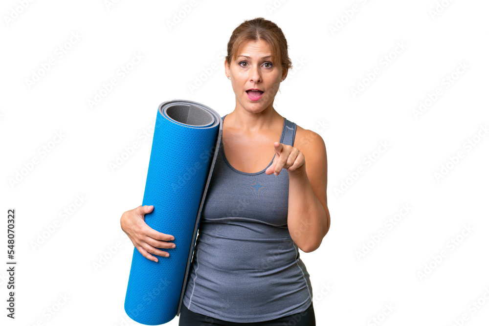 Middle-aged sport woman going to yoga classes while holding a mat over isolated background surprised and pointing front Middle-aged sport woman going to yoga classes while holding a mat over isolated background surprised and pointing front