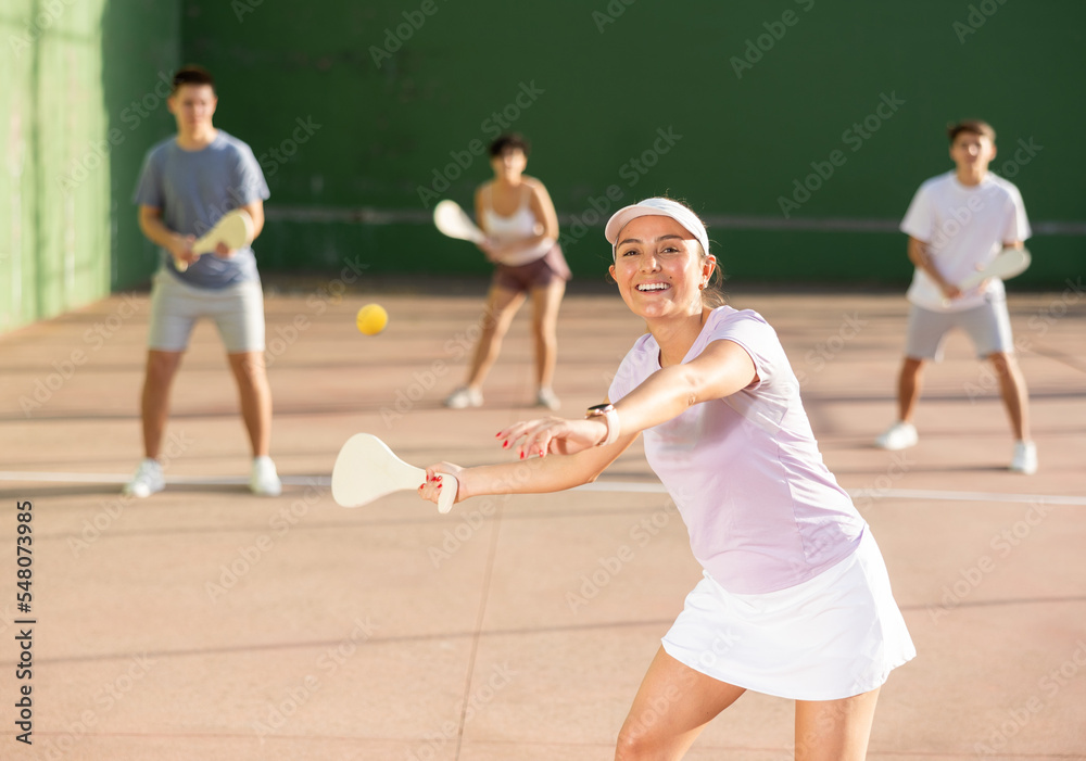 Concentrated young hispanic female pelota player swinging wooden paleta to hit ball on outdoor fronton court on sunny summer day. Sport and active lifestyle concept