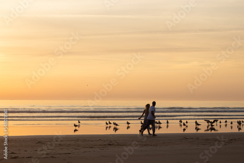 Warm couple walking on beach at sunset. Man and woman in casual clothes running along water at dusk. Love, family, nature concept